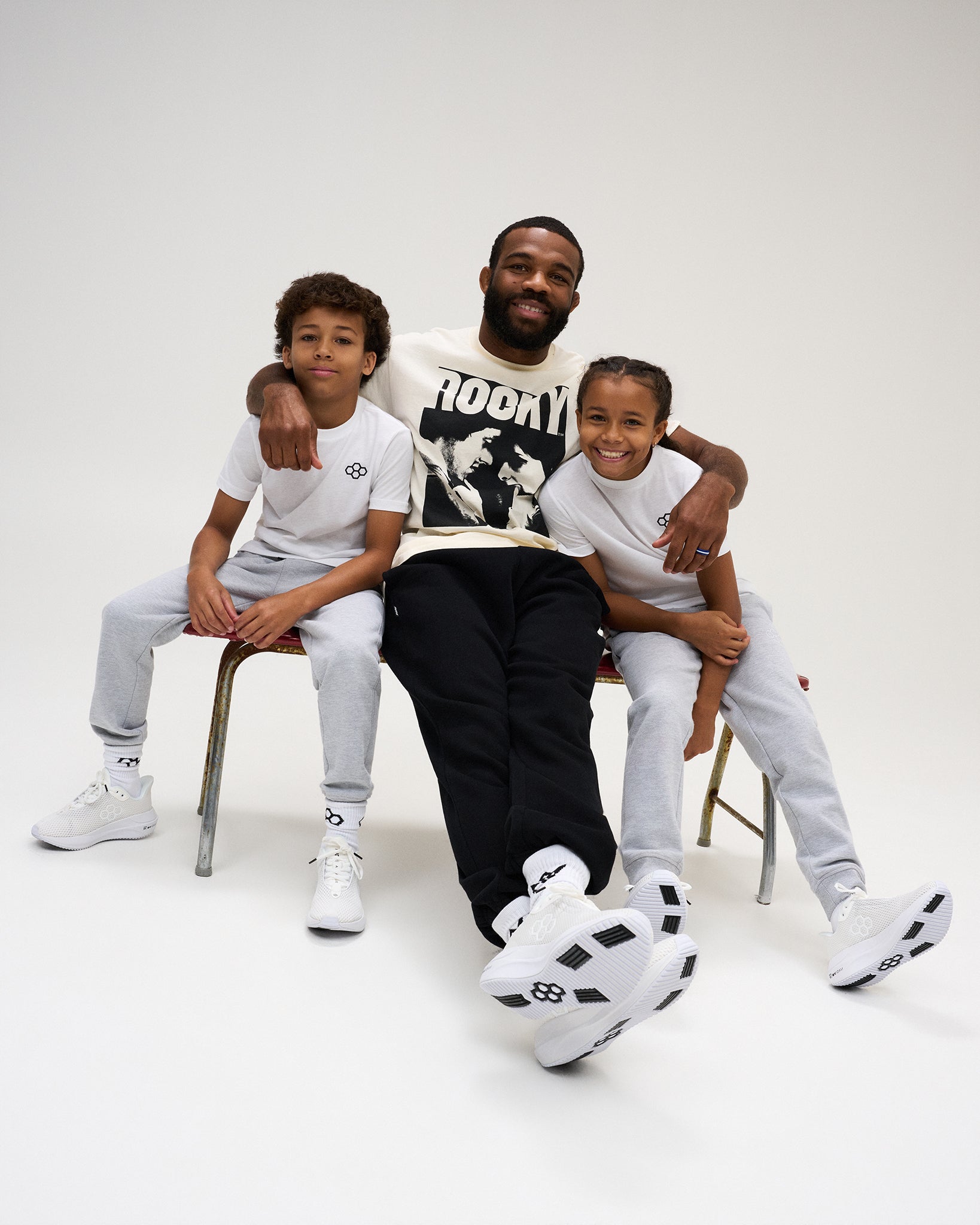 Two youth athletes wear white RUDIS Essential Hex Youth T-Shirts and gray joggers while seated with an adult in a studio setting.