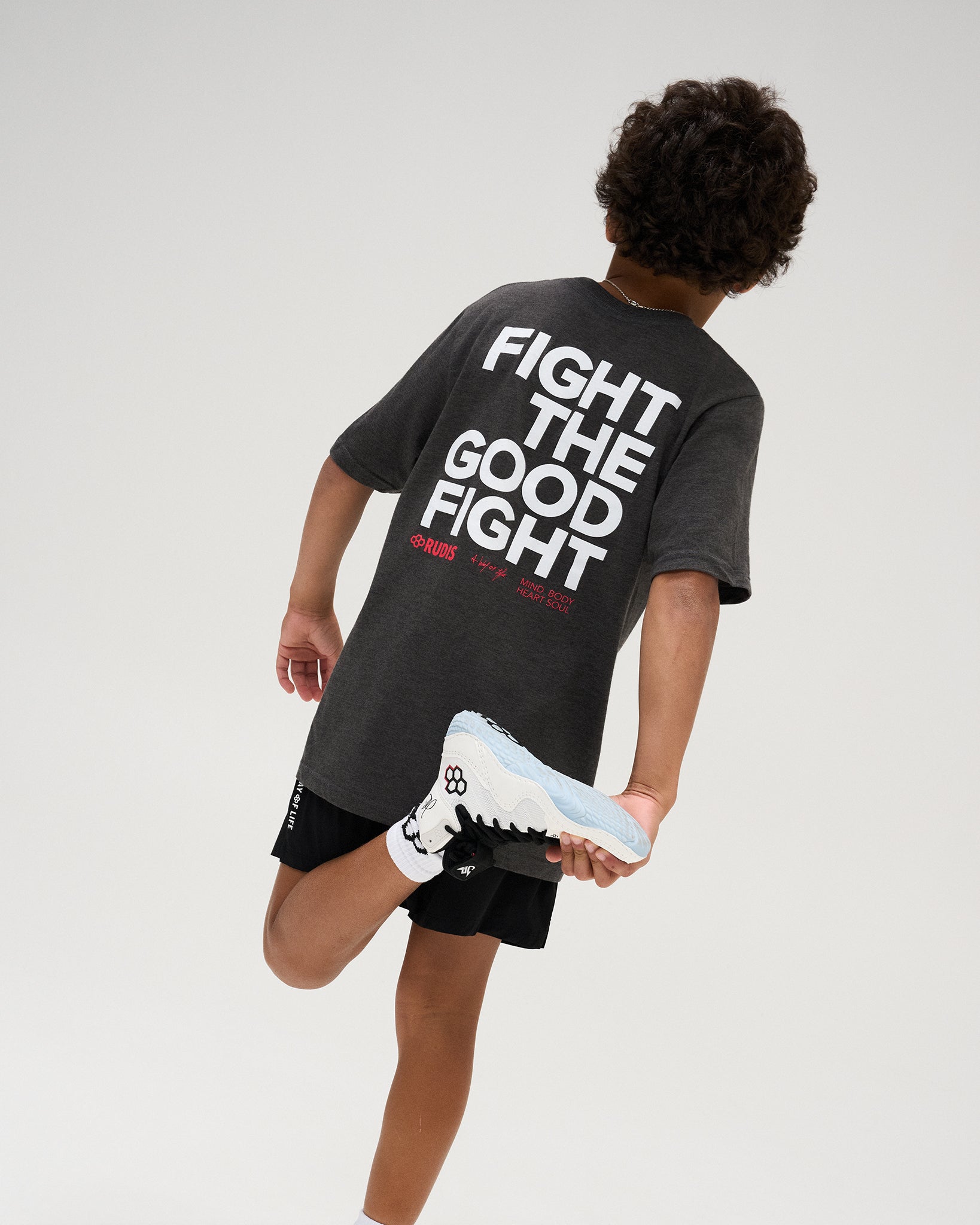 Youth athlete models the back of the charcoal RUDIS Fight the Good Fight Youth Heavyweight T-Shirt, featuring bold white and red graphics with black athletic shorts.