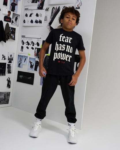 A child model wearing a black graphic t-shirt with the text 'fear has no power' in white gothic font, styled with black sweatpants and white sneakers, standing in a studio with fashion concept boards on the wall.
