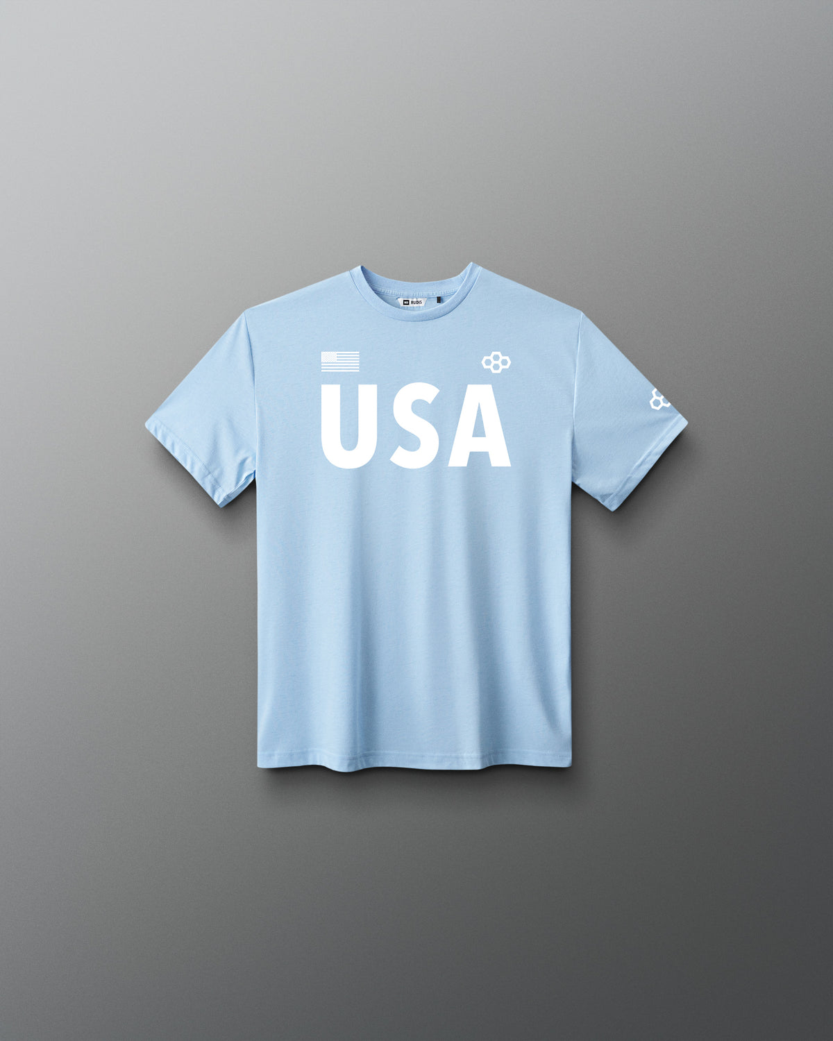 Front view of RUDIS USA Youth Elite Super Soft T-Shirt in light blue, featuring white USA lettering, an American flag graphic, RUDIS Hex logos, and short sleeves.