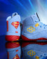 Close-up of RUDIS JB1 Superman wrestling shoes featuring light blue uppers, red soles, yellow RUDIS branding and Superman shield graphics.