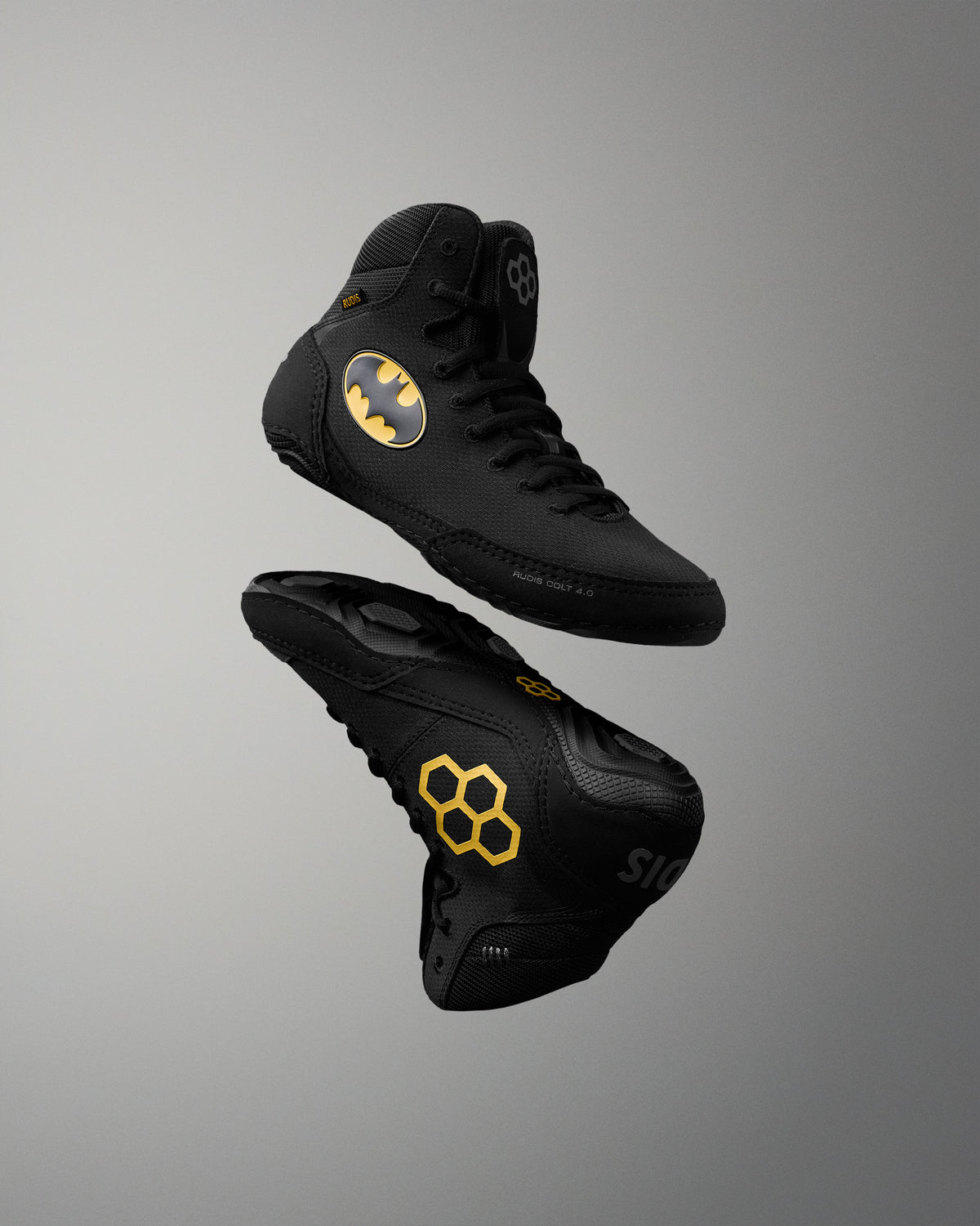 Two black high-top wrestling shoes floating against a gray gradient background. One shoe displays a yellow Batman logo on the side, and the other shows a yellow honeycomb pattern on the sole.