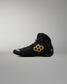 A black high-top wrestling shoe with a yellow geometric honeycomb logo on the side and a small gray logo on the heel, shown in profile against a neutral gray background.