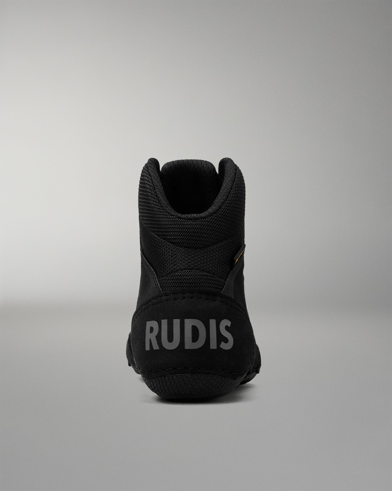A black mid-top wrestling shoe shown from the rear, featuring the brand name 'RUDIS' in gray lettering on the heel, set against a neutral gray studio background.