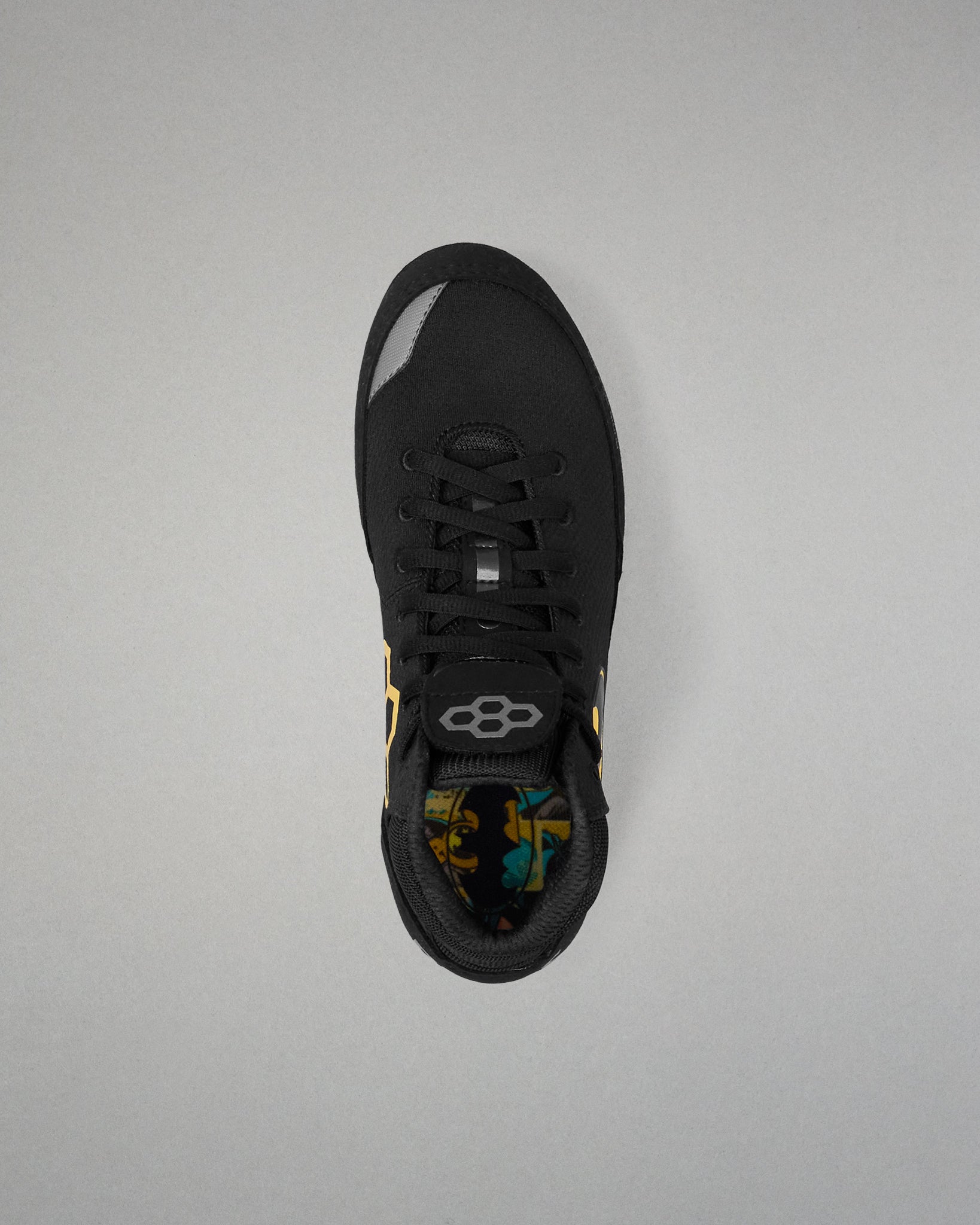 A black athletic wrestling shoe shown from a top-down view against a neutral gray background. The shoe features a black mesh upper, black laces, a black heel counter, and a small yellow accent on the side.