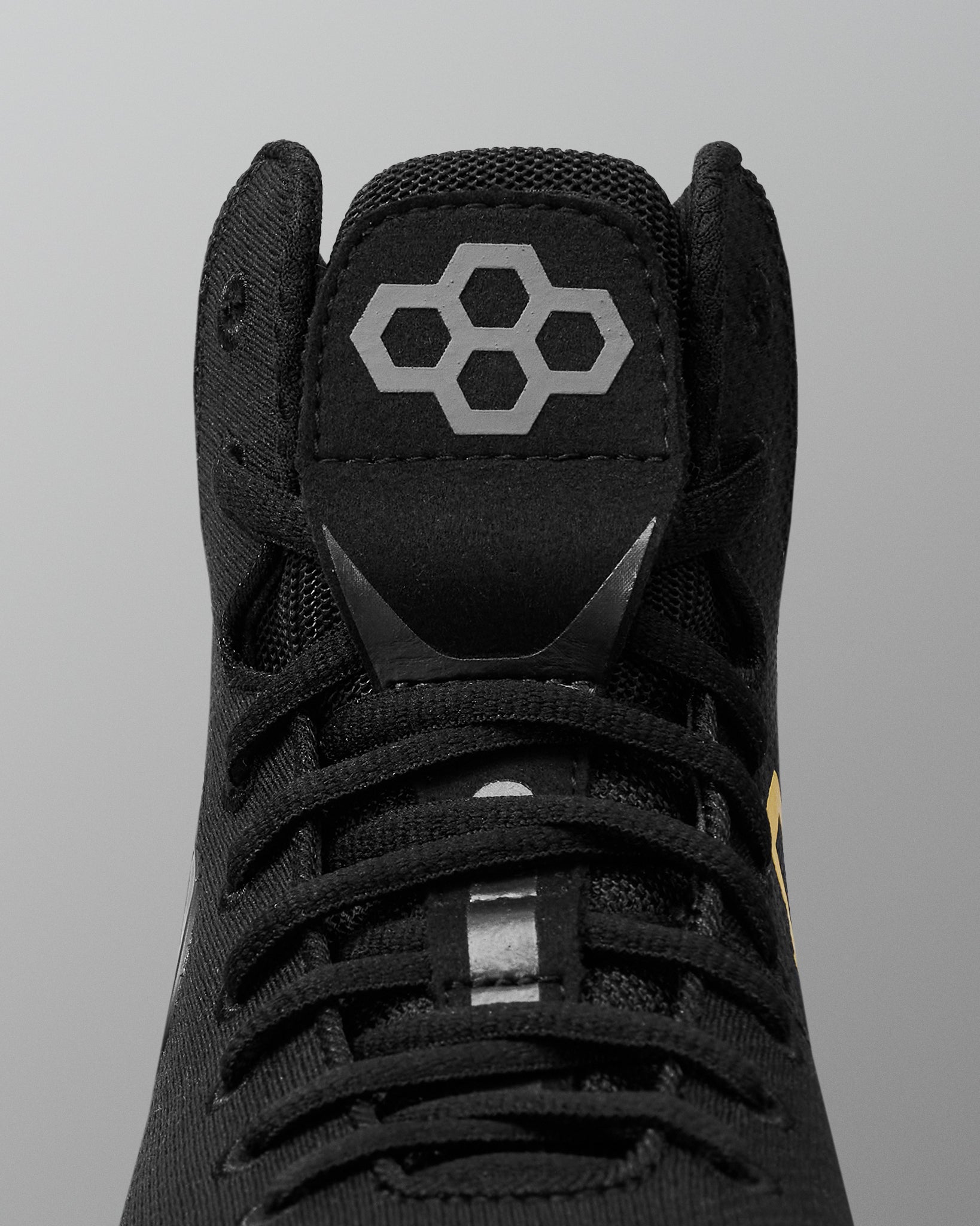 Close-up of a black high-top wrestling shoe showing the tongue with a gray geometric hexagon logo, black laces, and a small yellow accent near the side, set against a neutral gray background.