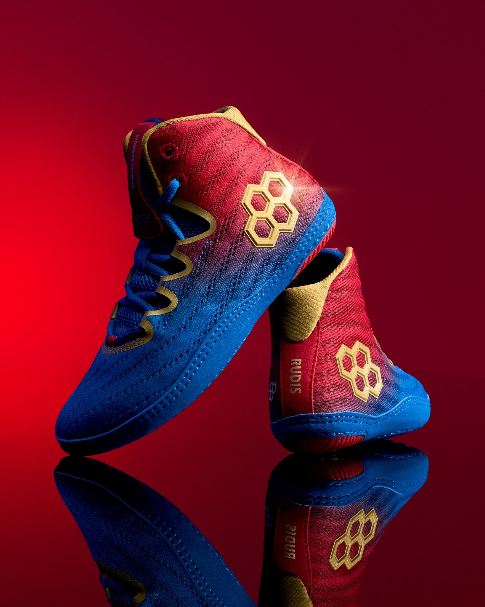 RUDIS Wonder Woman Hildebrandt 2.0 girls' wrestling shoes displayed against a red background, featuring red and blue uppers, gold logos and reflective styling.